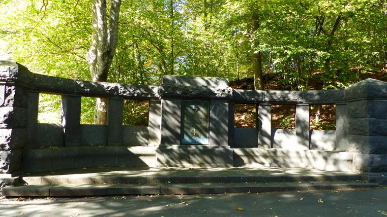 Ein steinernes Denkmal in einem grünen Wald. Es ist von Bäumen umgeben und der Boden ist mit Schatten bedeckt.