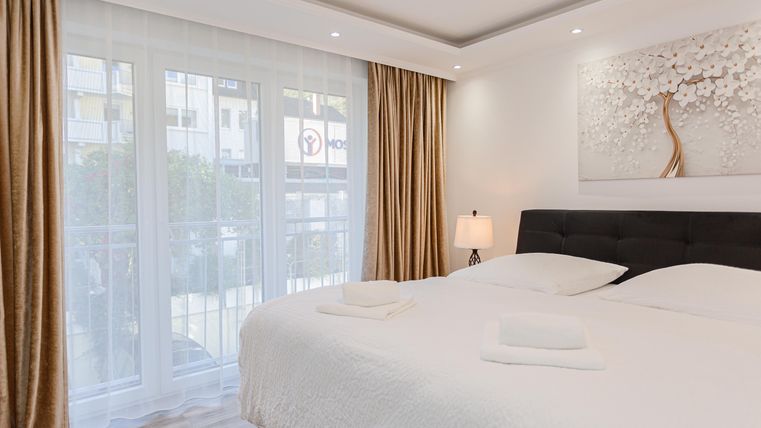 A modern bedroom with a large bed and a bedside table. Curtains and windows create a bright atmosphere.