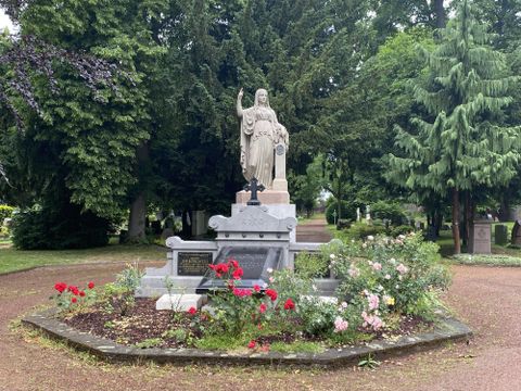 Een standbeeld staat in het midden van een vriendelijk aangelegd graf, omringd door fleurige bloemen. Op de achtergrond zijn hoge bomen zichtbaar die een rustige sfeer creëren.
