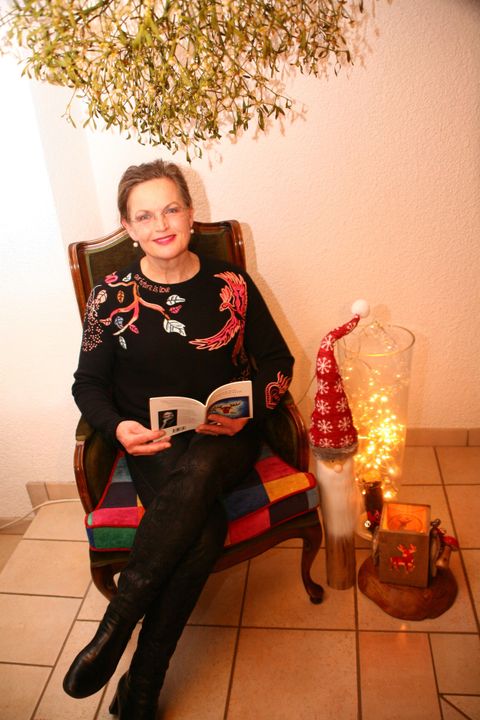 Eine Frau sitzt gemütlich in einem Stuhl und liest ein Buch. Neben ihr steht eine dekorative Weihnachtslaterne und ein kleiner Weihnachtsbaum.