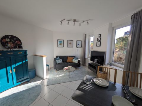 Ein helles Wohnzimmer mit einem blauen Schrank, einer gemütlichen Couch und einem Fernseher. Helle Wände und Fenster lassen viel Licht hinein.