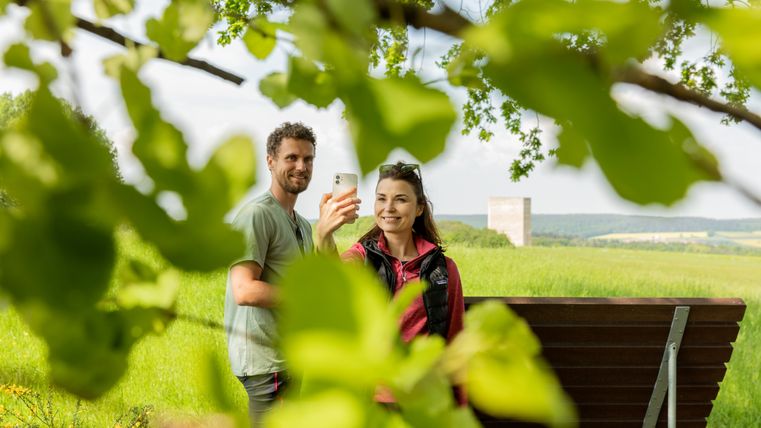 Twee mensen nemen een selfie voor de Bruder Klauskapel, omringd door groene bladeren.