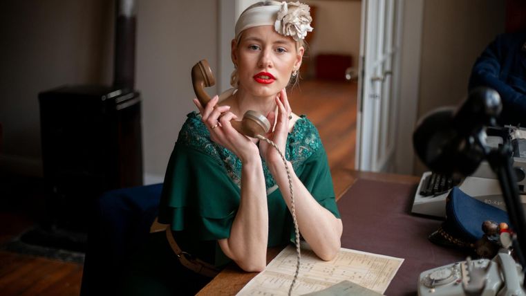 Une femme élégante avec du rouge à lèvres rouge est assise à un bureau et tient un combiné téléphonique. Elle porte une robe verte et a des fleurs dans les cheveux.