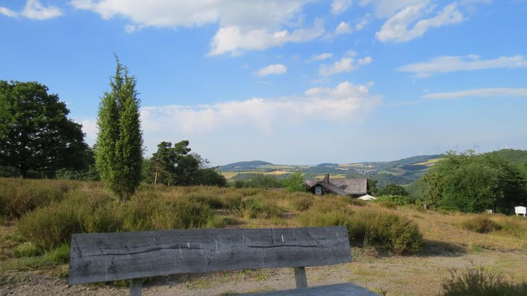Eine Bank steht in einer malerischen Landschaft mit sanften Hügeln und Bäumen. Der Himmel ist klar mit einigen Wolken.