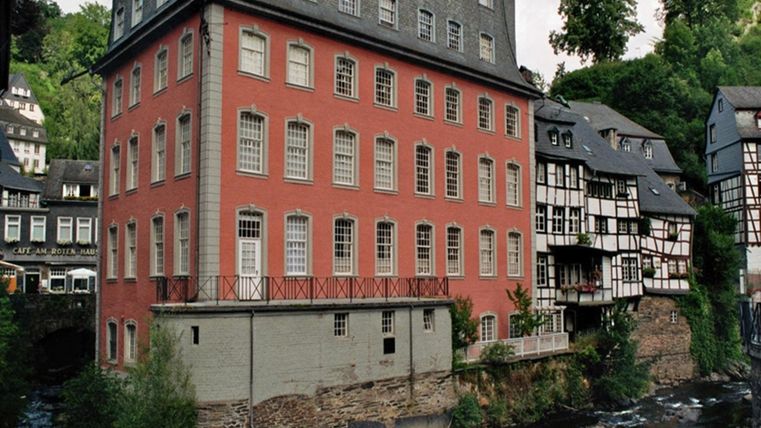 A red building next to a small river surrounded by traditional half-timbered houses. The scenery is green and picturesque.