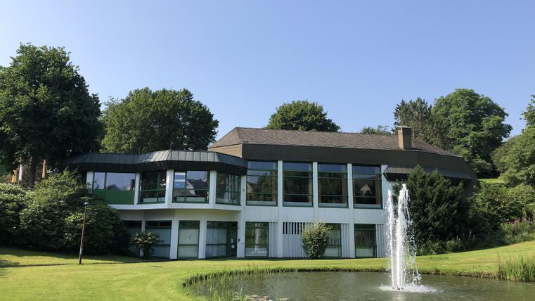 Ein modernes Haus mit großen Fenstern und einem gepflegten Garten. Im Vordergrund befindet sich ein Teich mit einem Wasserfall.