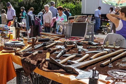 Ein Flohmarkt mit vielen alten Werkzeugen und Waffen auf einem Tisch. Im Hintergrund sind Menschen zu sehen, die die Stände anschauen.
