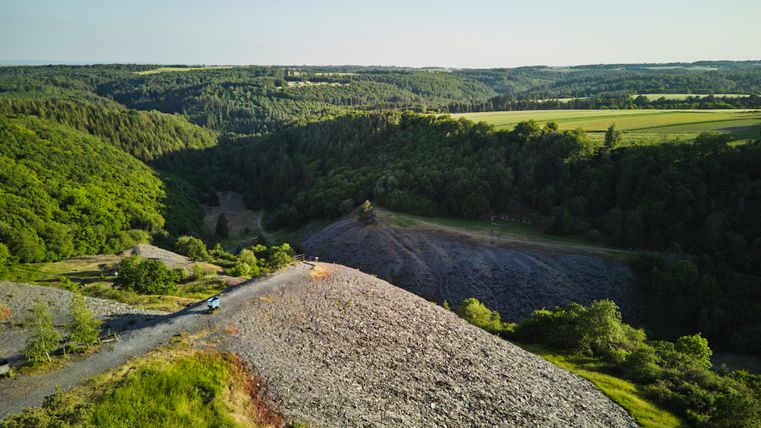Uitzicht over de Kaulenbachvallei in de Eifel: op de voorgrond een groene weide, daarnaast een grijs leisteenveld. Daarachter strekken beboste heuvels en dalen zich uit tot aan de horizon onder een blauwe hemel.