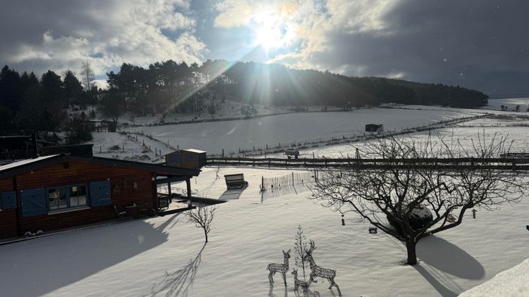 A winter landscape with lots of snow and bright sunlight. In the foreground, there is a wooden house and some decorative figures.