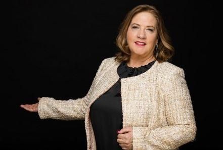 A woman in an elegant blazer stands in front of a black background. She is smiling and holding out a hand as if she is presenting something.