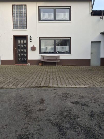 Une maison à deux étages avec une façade lumineuse. Devant la maison se trouve une zone pavée et un banc.