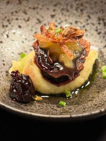Een kunstzinnig opgemaakt bord met gepureerde groenten, een stuk vlees en een fruitige saus. Het gerecht is gegarneerd met gefrituurde uien en verse kruiden.