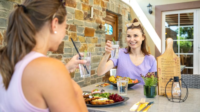Twee vrouwen proosten met een drankje aan een tafel buiten.
