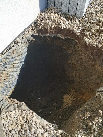 Un grand trou dans le sol, entouré de terre et de petits cailloux. Sur le mur, un filet est visible, qui sert probablement à des fins de drainage.