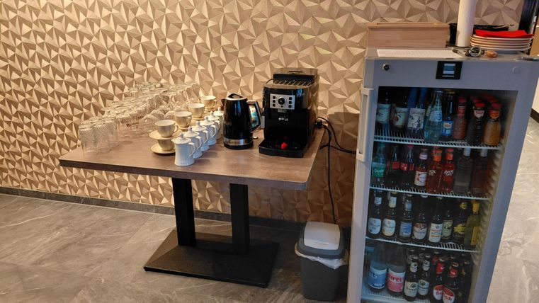 Une station de café avec des verres et une machine à café se trouve sur une table. À côté, il y a un réfrigérateur avec différentes boissons.