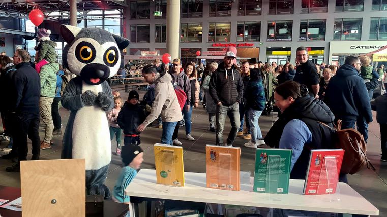 Een gigantisch dierenkostuum staat aan een tafel met meerdere kleurrijke boeken. Op de achtergrond is een menigte te zien in een drukke omgeving.