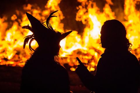 Zwei Silhouetten stehen vor einem großen Feuer. Eine Person trägt einen auffälligen Hut, während die andere spricht.