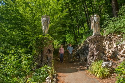 Two people are walking along a forest path between two statues of angels.
