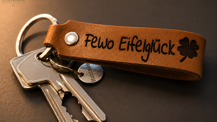 Ein Schlüsselbund mit einem braunen Lederanhänger, auf dem "Fewo Eifelglück" eingraviert ist. Zusätzlich sind einige Schlüssel sichtbar, die daran hängen.