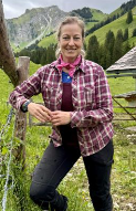 Eine Frau steht an einem Zaun in einer schönen Berglandschaft. Im Hintergrund sind grüne Wiesen und hohe Berge zu sehen.