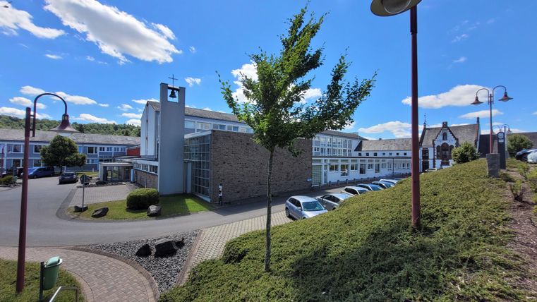 Ein modernes Gebäude mit einer skylight und grünen Bäumen vor einem blauen Himmel. Parkplatz mit mehreren Autos zu sehen.